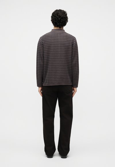 Άνδρας με σγουρά μαλλιά στέκεται με την πλάτη προς τα εμπρός, φορώντας ένα σκούρο γκρι πουκάμισο με σχέδια και μακριά μανίκια και φαρδιά μαύρα παντελόνια μπροστά σε απλό λευκό φόντο.