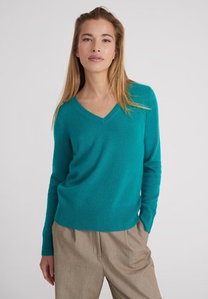Sukienka w kolorze teal z dekoltem w serek, wykonana z miękkiego materiału, z prążkowanymi mankietami i dołem, w połączeniu z jasnobrązowymi spodniami, tworząc kontrast kolorów i faktur.