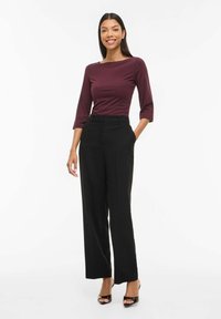 Smaidīga sieviete, tērpusies burgundijas krāsas trīs ceturtdaļu piedurkņu topā, ar augstu jostas vietu melnās plaša piegriezuma biksēs un melnās atklātās purngalu kurpēs, stāv ar vienu roku kabatā.