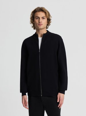 Giovane uomo con capelli biondi ondulati che indossa un maglione nero con cerniera sopra una camicia bianca, in piedi davanti a uno sfondo chiaro e uniforme.