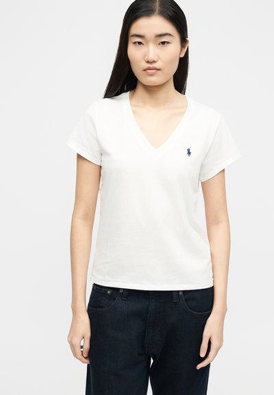 Γυναίκα με μακριά μαύρα μαλλιά που φοράει λευκό μπλουζάκι V-neck με κοντά μανίκια και μικρό ναυτικό λογότυπο, καθώς και σκούρο μπλε τζιν, στέκεται μπροστά σε λευκό φόντο.