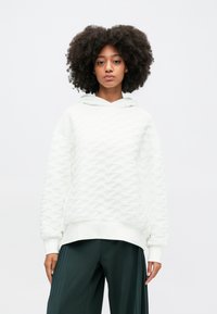 Biała teksturowana bluza z kapturem w falistym wzorze, z kapturem i ściągaczami przy mankietach, połączona z ciemnozielonymi spodniami o szerokich nogawkach.