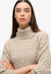 Suéter de punto beige con un alto cuello acanalado y patrón de trenzas, con mangas largas y un corte relajado. Textura suave y material ligero.