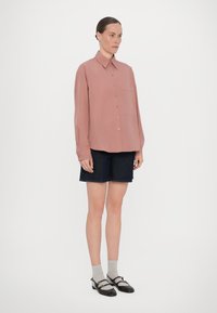 Helles rosa Langarmhemd mit Kragen und Brusttasche, kombiniert mit dunkelblauen Jeansshorts. Modell trägt graue Socken und schwarze Ballerinas.