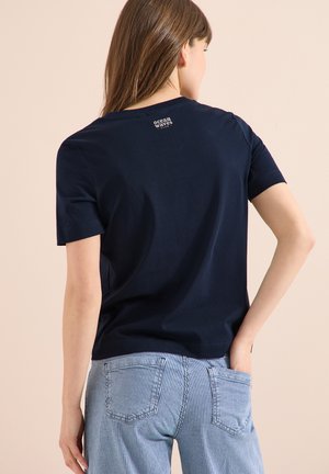 Femme aux longs cheveux bruns portant un t-shirt à manches courtes bleu marine et un pantalon à rayures bleu clair, une main dans la poche, vue de dos.