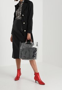 Genomskinlig tote-väska med svart inredning, hålls av en person i svart kappa, leopardmönstrad skjorta och röda ankelstövlar.