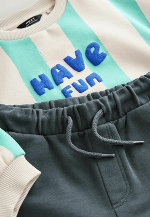 Licht beige sweatshirt met turquoise strepen en blauwe gestructureerde tekst "HAVE FUN" bovenop donkere grijze joggingbroek met elastische tailleband en koord.