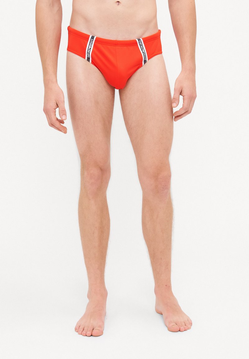 Rote Schwimmslips aus dehnbarem Material, mit einem hochgeschnittenen Design und schwarz-weißen gestreiften Akzenten am Bund.