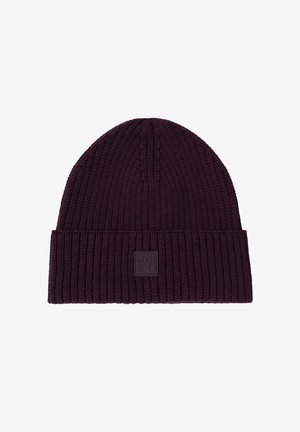 Femme portant un manteau matelassé bleu foncé, un pull rose, un jean à jambes larges, des baskets blanches et un bonnet tricoté violet foncé, debout et souriante.