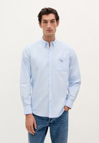 Lichtblauwe overhemdd met een borstzak, voorzien van een klein geborduurd ontwerp. Gemaakt van een soepele stof met een kraag.