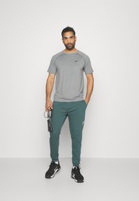Grå trenings T-skjorte med svart Nike-logo, parret med grønne sweatpants med logoakenter. Joggesko med svart og hvit detalj, holder et hoppetau.