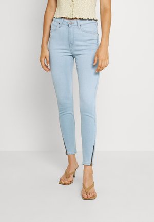 Ljusblå skinny jeans med hög midja, utrustade med sidodragkedjor vid anklarna och en mjuk denimtextur. Matchas med en blommig crop top.