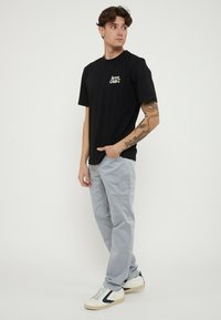 Joven con el brazo tatuado, lleva una camiseta negra con gráfico, pantalones gris claro y zapatillas blancas, está de pie con una mano en el bolsillo y mira de lado.