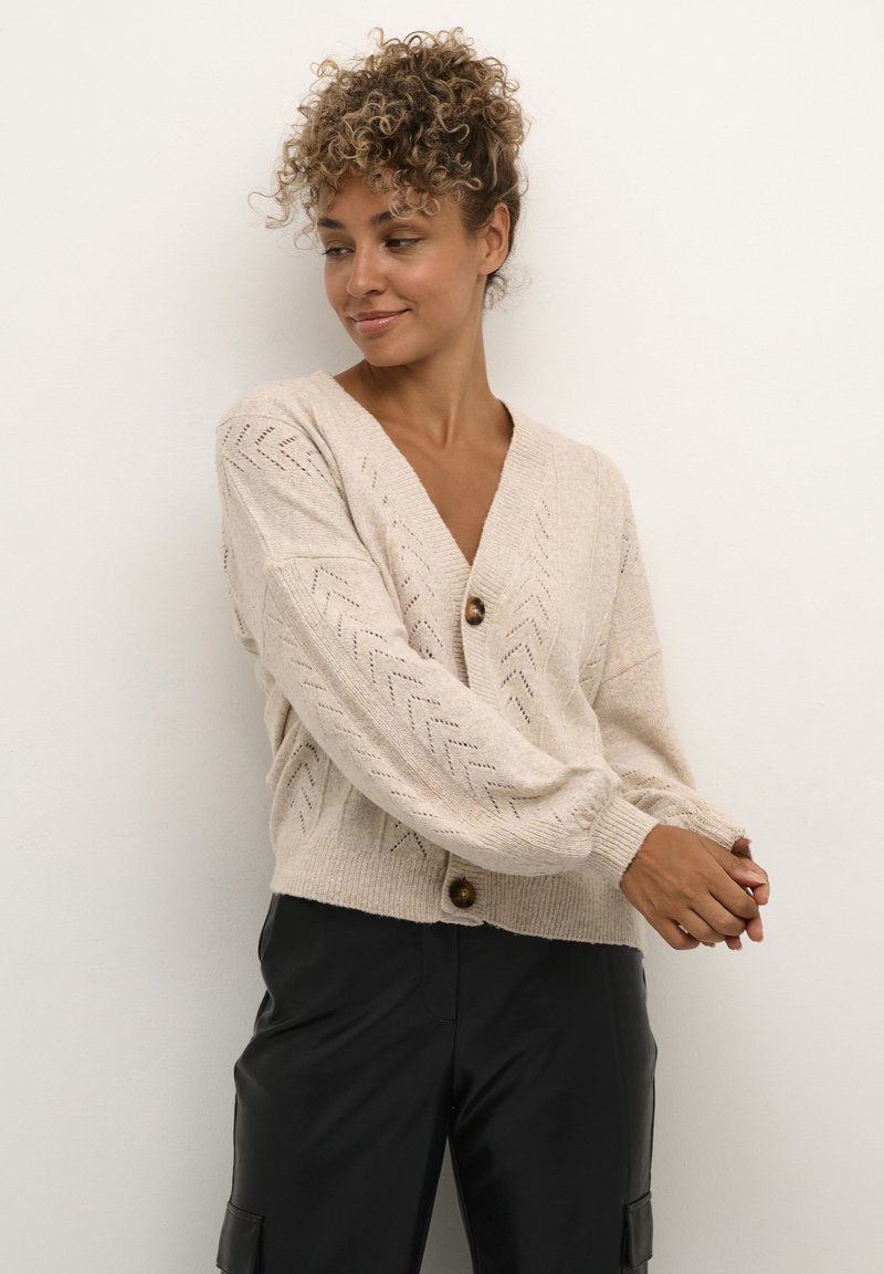 Beige strikket cardigan med dyb V-hals, zigzag mønster, lange ærmer og en enkelt lukning med træknap. Blødt, tekstureret stof.