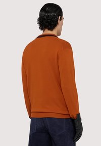 Pomarańczowy sweter z kołnierzykiem, z długimi rękawami i ściągaczami. W połączeniu z ciemnoniebieskimi spodniami i czarnymi skórzanymi rękawiczkami.