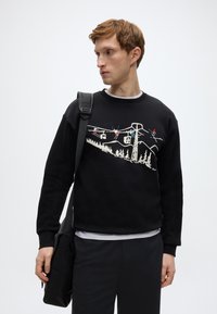 Μαύρο φούτερ με γραφικό σχέδιο που απεικονίζει σκιέρ και τελεφερίκ με φόντο ένα ορεινό τοπίο. Μαλακό ύφασμα και στρογγυλή λαιμόκοψη.