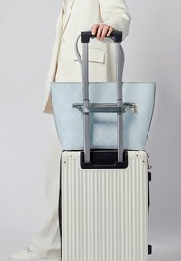 Borsa a tracolla azzurro chiaro con un motivo geometrico e cerniera argentata, appoggiata su una valigia bianca con coste e manico telescopico.