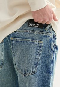 Svijetloplave traperice s istrošenim izgledom, sa stražnjim džepovima i crnom tkaninom oznake s natpisom "DENIM TOM TAILOR."