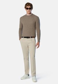Brauner Kabelstrickpullover, helle Hosen, schwarzer Gürtel und weiße Sneakers mit schwarzen Akzenten. Modell trägt Sonnenbrille und posiert aufrecht.