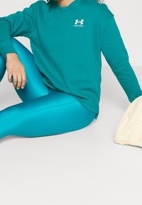 Miękka, turkusowa bluza z białym logo, zestawiona z błyszczącymi, turkusowymi legginsami. Delikatny materiał i luźny krój, obok kremowa torba.