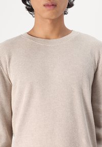 Chemise à manches longues en tricot gaufré beige avec un col rond. Tissu texturé et coupe ample, avec des coutures visibles le long de l'encolure et des manches.