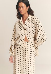 Cream-colored button-up shirt with black polka dots, featuring a relaxed fit, long sleeves, and a cropped design. Paired with matching polka dot skirt.