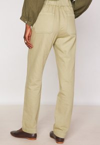 Vrouw in hooggetailleerde beige broek met achterzakken, gecombineerd met donkerbruine flatjes en een olijfgroen shirt met lange mouwen.