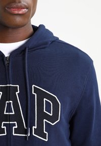 Marinblå zip-hoodie i bomull med en stor vit "GAP"-logotyp i strukturerad tyg på vänster bröst.