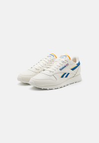 Białe sneakersy z niebieskimi i żółtymi akcentami, skórzany wierzch o fakturze, sznurowana konstrukcja oraz gruba podeszwa z bieżnikiem. Widoczne logo Reebok.