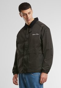 Black jacket with a corduroy collar, snap buttons, and embroidered logo on the left chest. Features elastic cuffs and two front pockets.