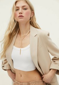 Bež blazer nad belim rebrastim crop topom, v kombinaciji z bež hlačami na vrvico. Zlata večplastna ogrlica poudarja dekolte.