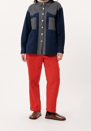 Personne portant une chemise bleu marine à boutons avec des rayures verticales, un pantalon rouge vif et des chaussures marron sans lacets, debout devant un fond uni.