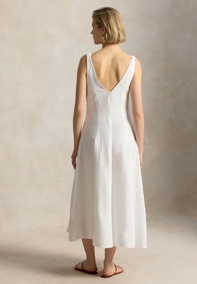 Sieviete stāv ar muguru pret mums, valkā baltu bezroku midi kleitu ar dziļu V izgriezumu mugurā un brūnas plakanas sandales.