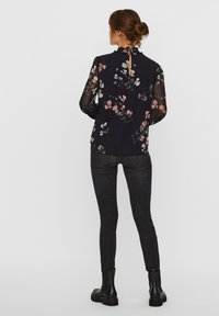 Blusa nera floreale con maniche lunghe trasparenti, scollatura arricciata e dettaglio a chiave sulla schiena. Abbinata a jeans scuri e stivaletti.