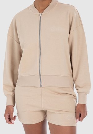 Beige Jacke mit Reißverschluss und passenden Shorts mit gerippten Bündchen und Bund, getragen von einer Person, mit dezent besticktem Text auf Brust und Shorts.