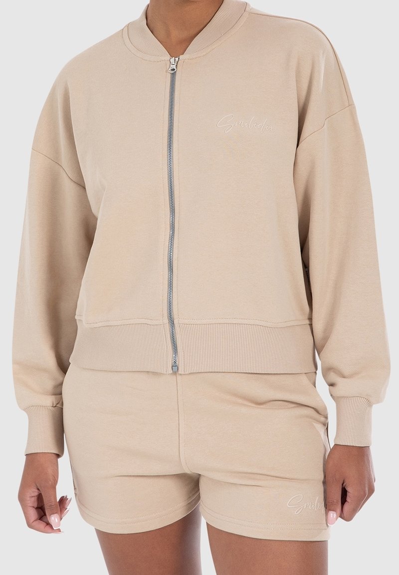 Veste beige zippée et short assorti avec poignets et taille côtelés portés par une personne, avec un texte brodé discret sur la poitrine et le short.