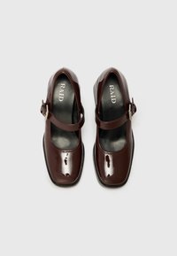 Chaussures Mary Jane en cuir verni bordeaux avec un bout rond, une bride ajustable et une semelle plate en caoutchouc. Logo visible sur la semelle intérieure.