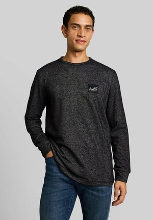 Homme portant une chemise à manches longues gris foncé avec un petit logo montagne sur la poitrine et un jean bleu, debout devant un fond gris clair.
