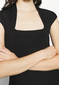 Mustat ribbed lyhythihaiset paidat, joissa on neliömainen kaula-aukko, näyttävät istuvalta ja sileältä. Käsi on asetettu kehon yli.