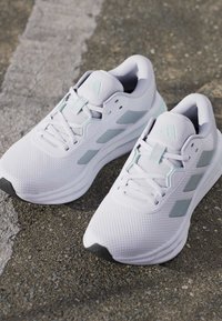 Paire de chaussures de course blanches et gris clair avec lacets posées sur l'asphalte près d'une ligne blanche délavée.