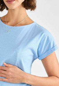 Ljusblå bomullströja med rullade korta ärmar. Mjuk textur och rund halsringning. Silverhalsband med två sammanflätade ringar.