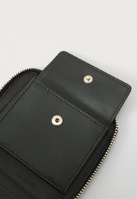 Portefeuille en cuir noir avec fermeture éclair, présentant une poche à rabat avec bouton-pression et des accents en métal doré. Texture lisse et lignes épurées.