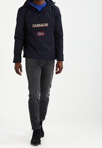 Marineblauwe pulloverjas met een voorzak, voorzien van een patch van de Noorse vlag en een logo. Gecombineerd met donkergrijze jeans en zwarte schoenen.