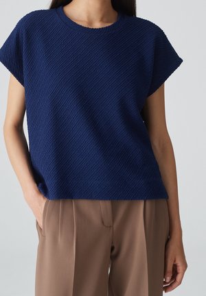 Marineblå struktureret strikbluse med korte ærmer, rund halsudskæring og diagonalt ribmønster, kombineret med brune, løstsiddende bukser.