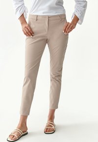 Pantaloni slim-fit beige con una texture liscia, dotati di chiusura con bottone frontale e tasche laterali. Indossati con sandali con lacci.