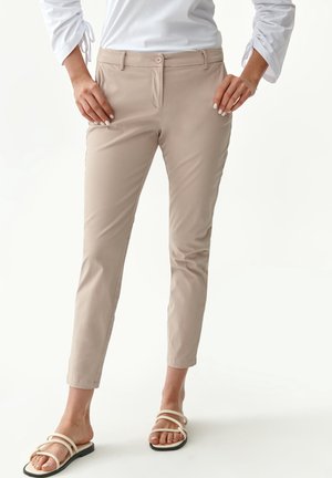 Pantalons slim beige avec une texture lisse, dotés d'une fermeture à bouton à l'avant et de poches latérales. Portés avec des sandales à lanières.