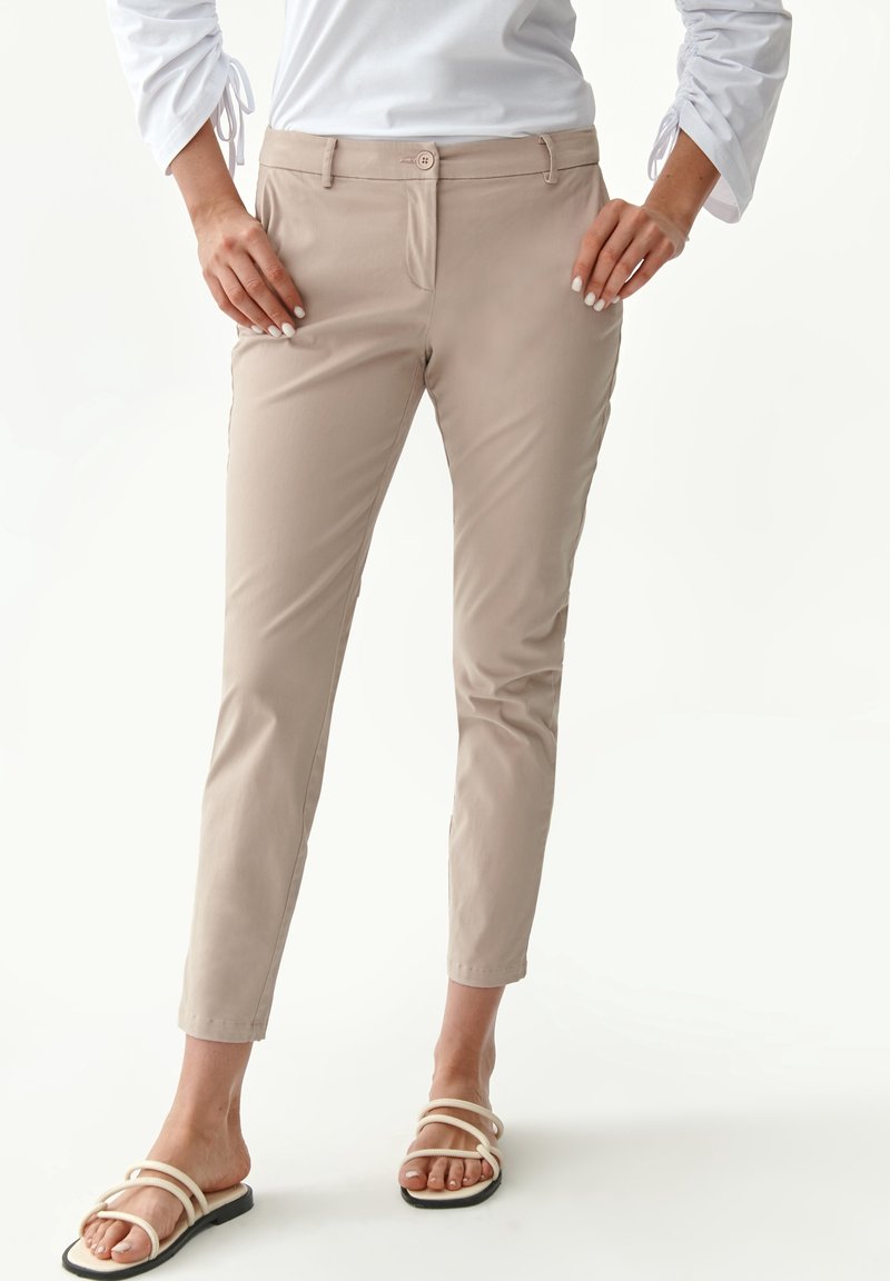 Pantaloni slim-fit beige con una texture liscia, dotati di chiusura con bottone frontale e tasche laterali. Indossati con sandali con lacci.