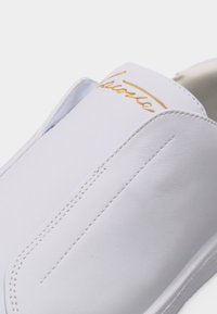 Κοντινή λήψη ενός λευκού δερμάτινου παπουτσιού χωρίς κορδόνια με ορατή ραφή και χρυσό καλλιγραφικό λογότυπο στην περιοχή της γλώσσας.