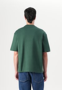 Tricou verde cu mânecă scurtă, cu o croială lejeră și decolteu tip crew, fabricat dintr-un material moale din bumbac, purtat cu blugi din denim albastru.