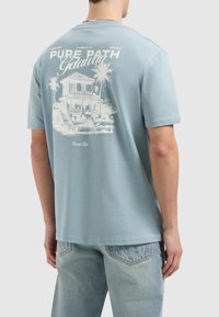 Lichtblauw katoenen t-shirt met een witte afbeelding van een huis en palmbomen op de achterkant. Relaxed fit met korte mouwen en een ronde hals.